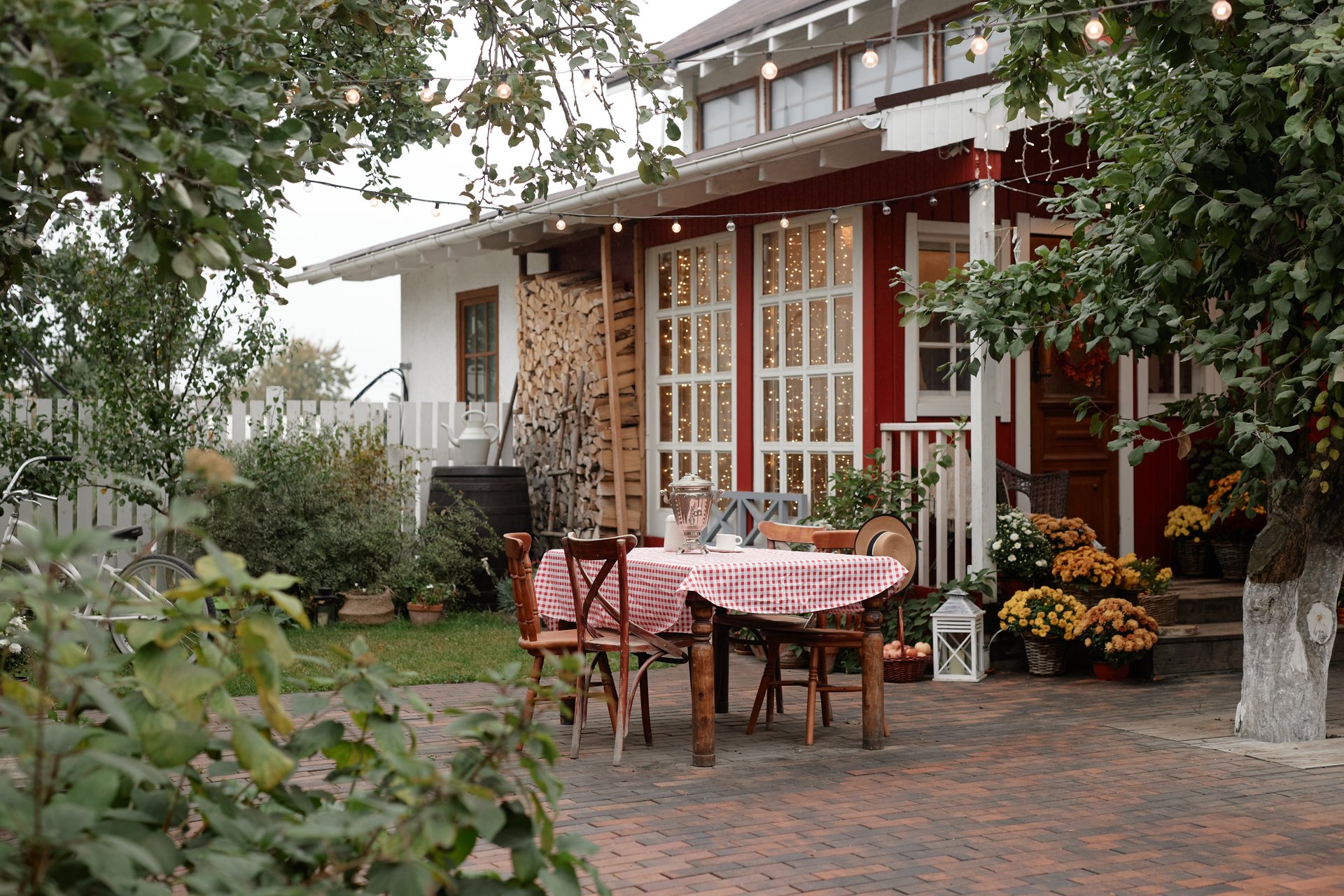 Empty outdoor table with checkered tablecloth standing in front of rustic village house surrounded by greenery and flowers, wooden chairs arranged around table, cozy rural setting