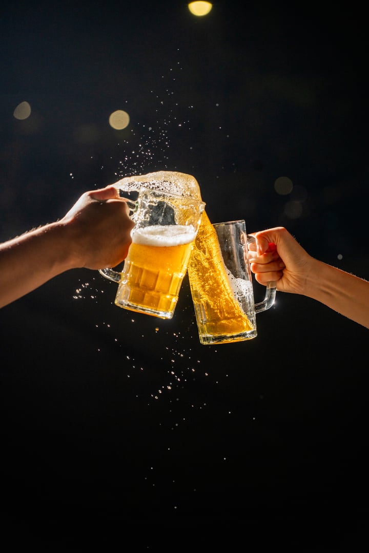 Two hands raise beer mugs overflowing with foam in a lively toast to celebrate Beer Fest. The beer spills from the mugs, adding to the festive atmosphere of this classic German festival
