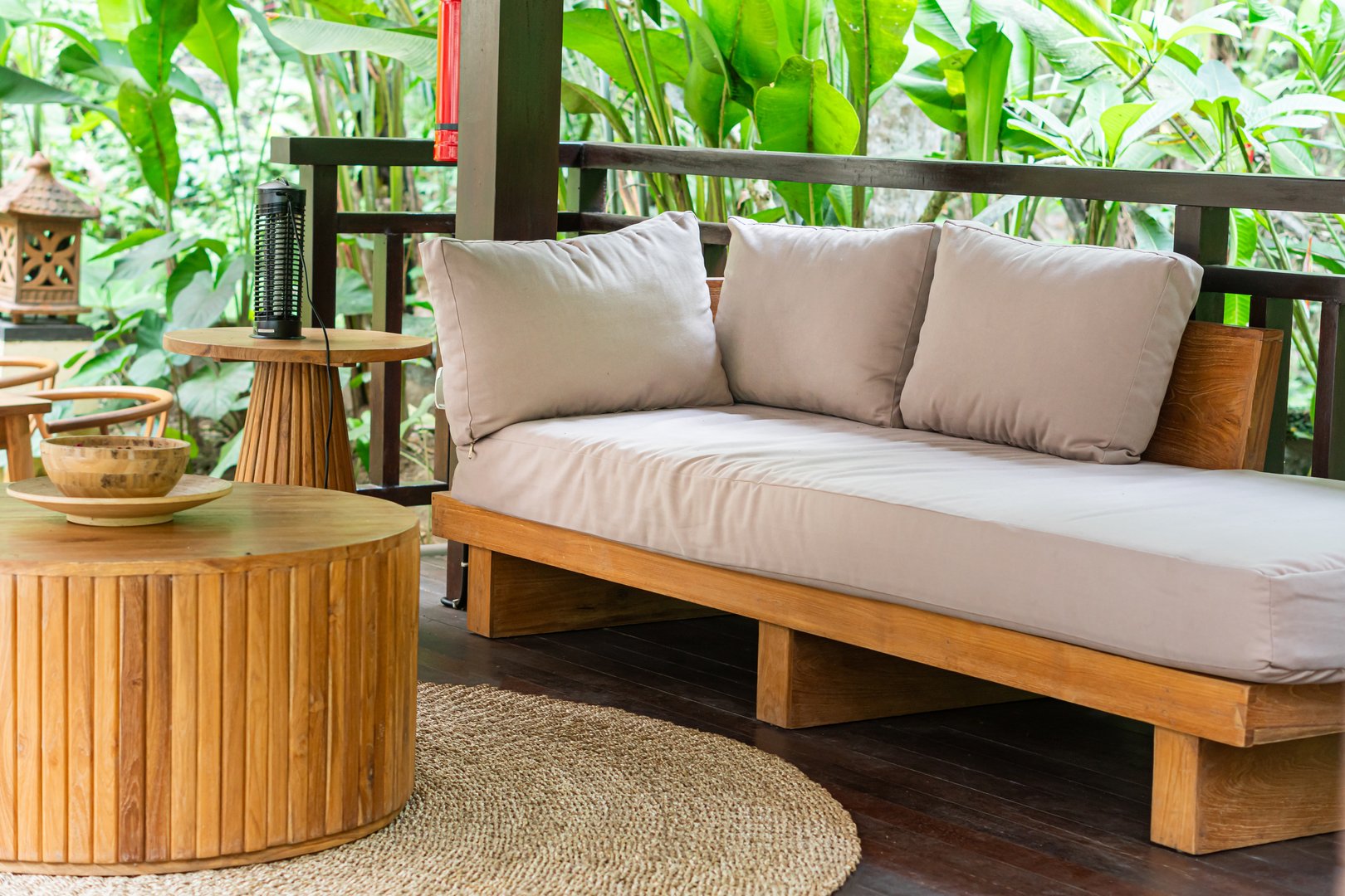 A cozy outdoor seating area featuring a wooden sofa with beige cushions, a round wooden table, and a woven rug. Lush green plants surround the space, creating a tranquil atmosphere.