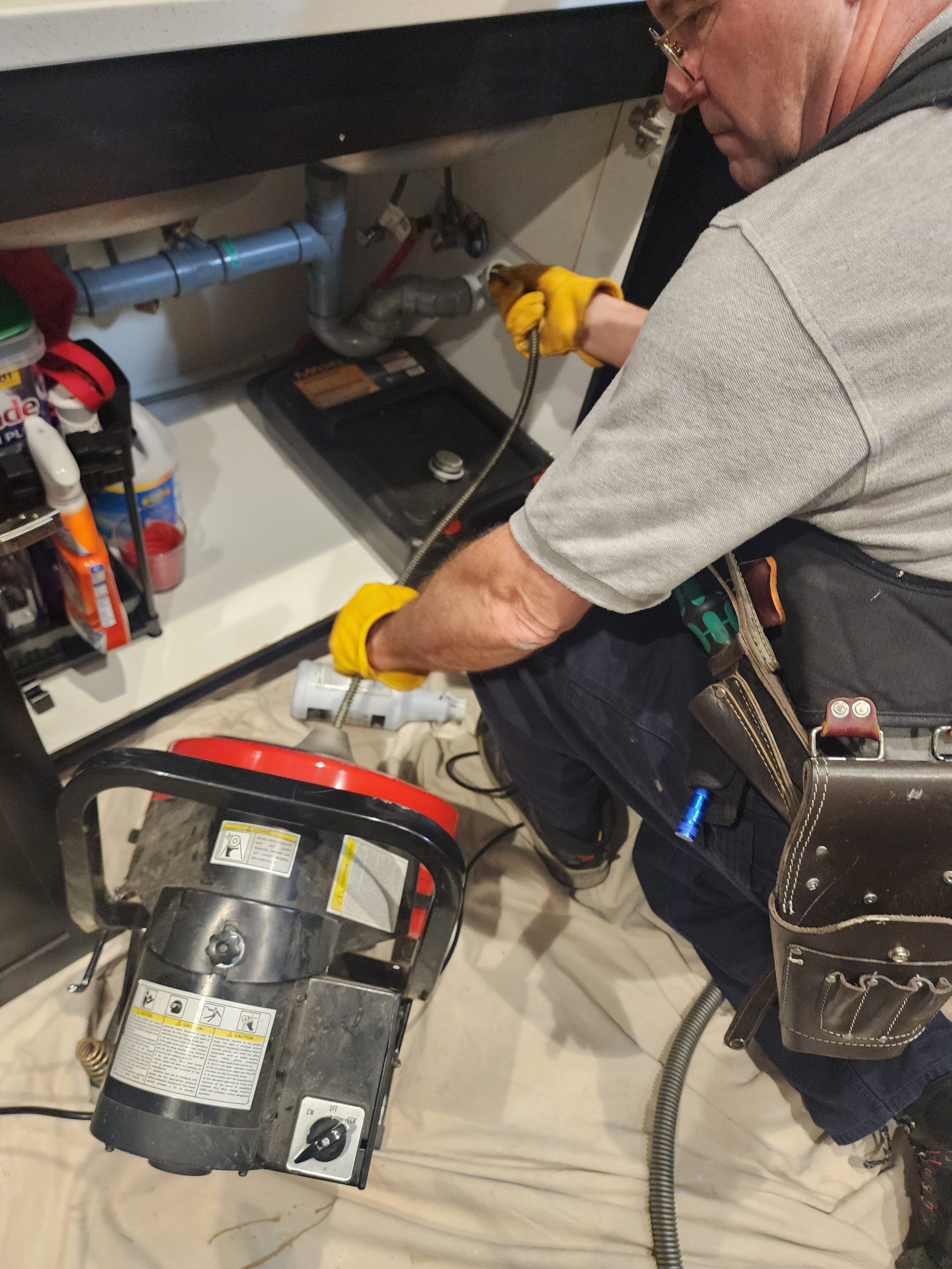 Plumber using a drain snake under a sink, with tools and cleaning supplies nearby.