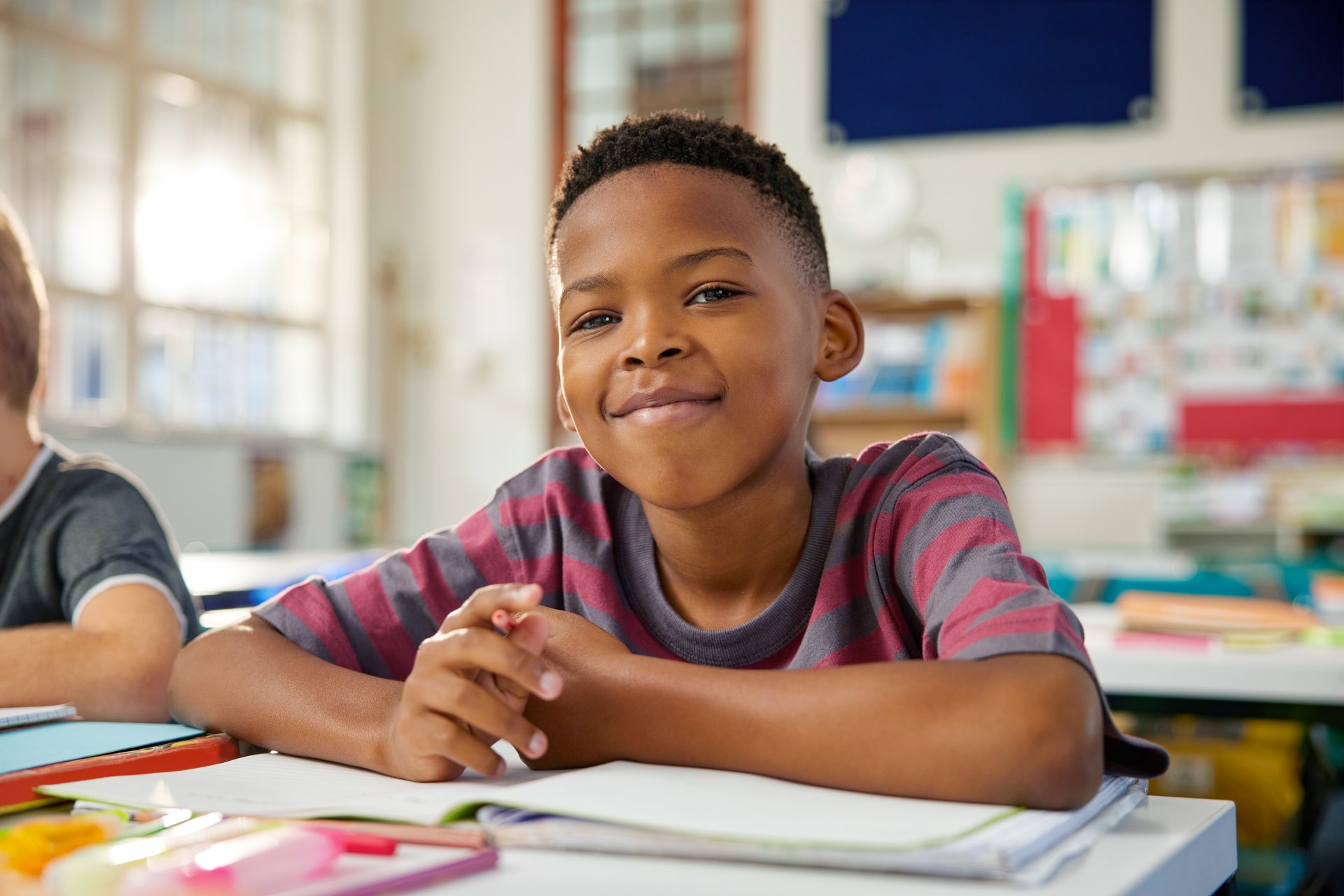 Portrait of happy black elementary boy studying in classroom while looking at camera. Close up face of little satisfied black boy writing in notebook using pencil. Proud schoolchild studying in classroom in primary school.