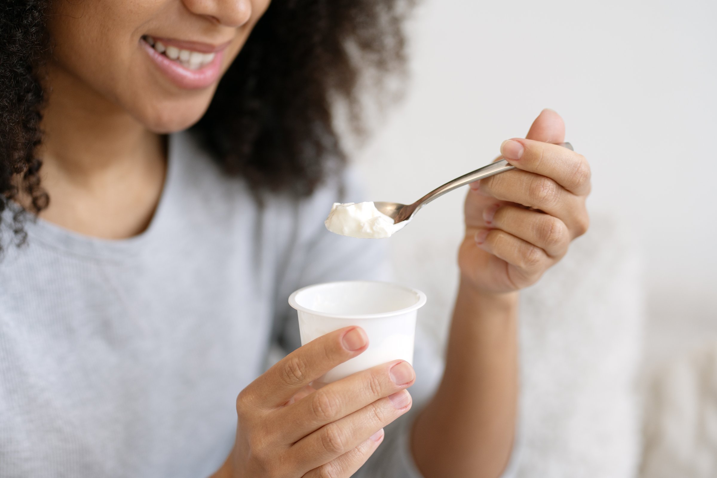 Smiling woman enjoying yogurt from a plastic cup, savoring each creamy spoonful while embracing a healthy lifestyle indoors