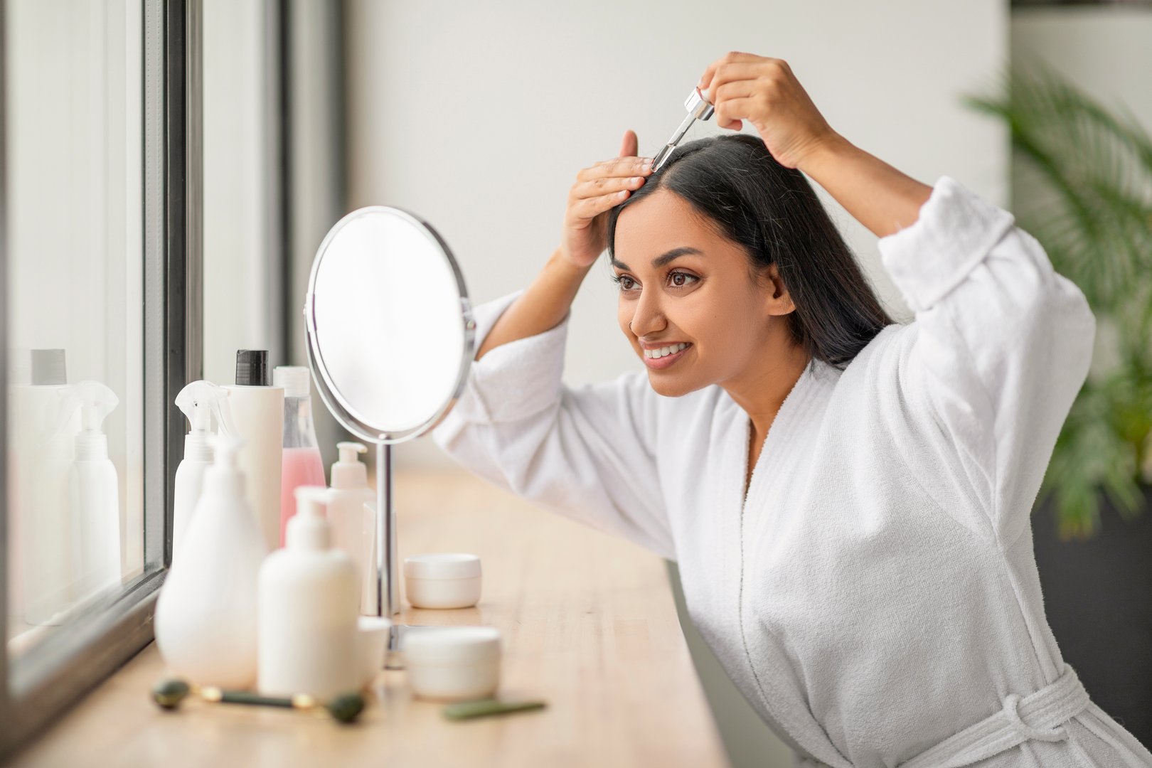 Positive pretty young indian woman wearing white bathrobe looking at mirror, using nourishing hair oil for hair growth, dropping serum on scalp. Hair care at home
