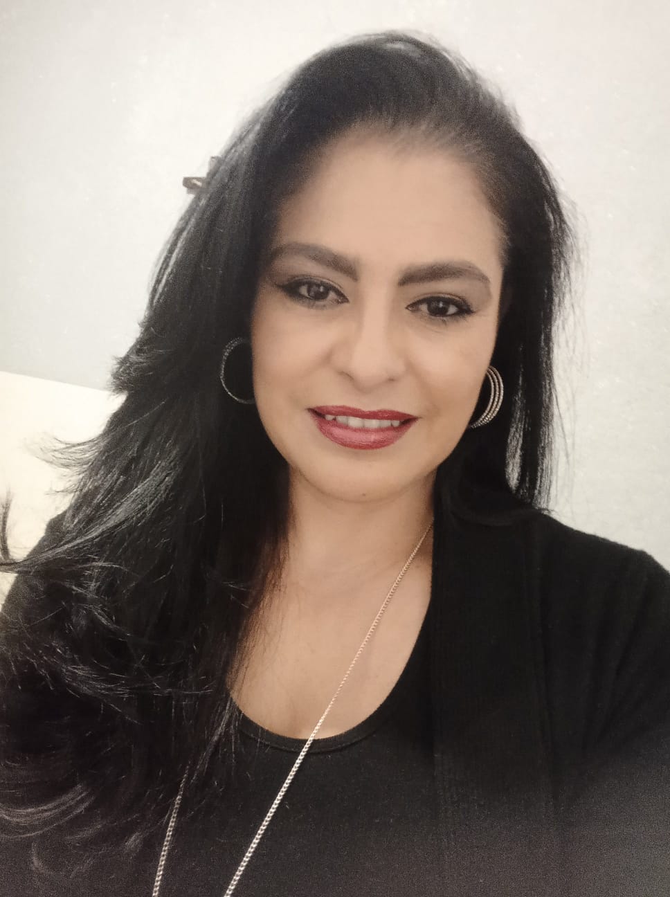 Woman with long dark hair, wearing hoop earrings and a necklace, smiling and seated against a light background.