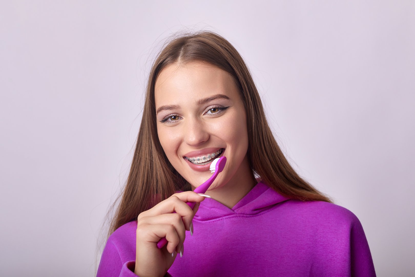 A young woman with braces on her teeth is brushing them with a toothbrush. She has a happy expression on her face and is looking at the camera. The background is a solid color.
