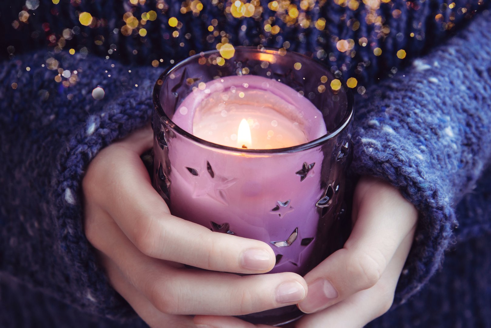 Girl in soft warm dark blue knitted sweater holds in hands burning candle with golden blur. Christmas candle. Waiting for the holiday