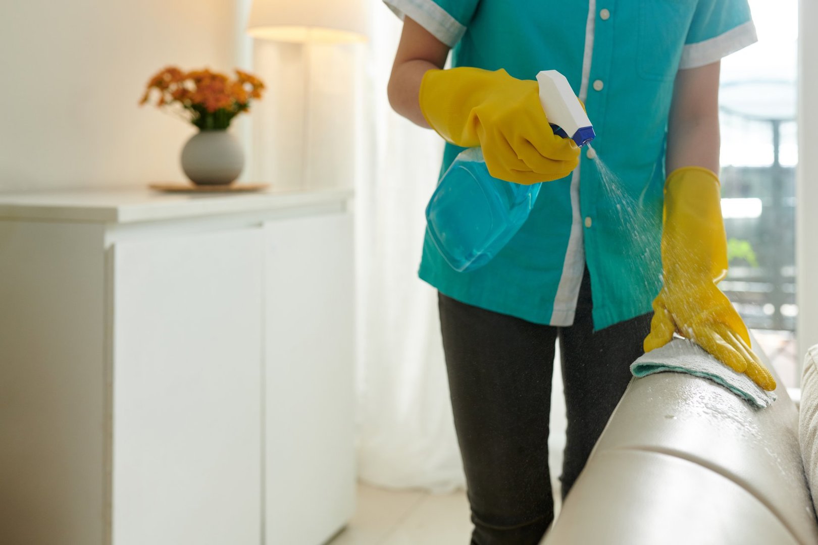 House cleaner in silicone gloves spraying detergent on beige leather couch