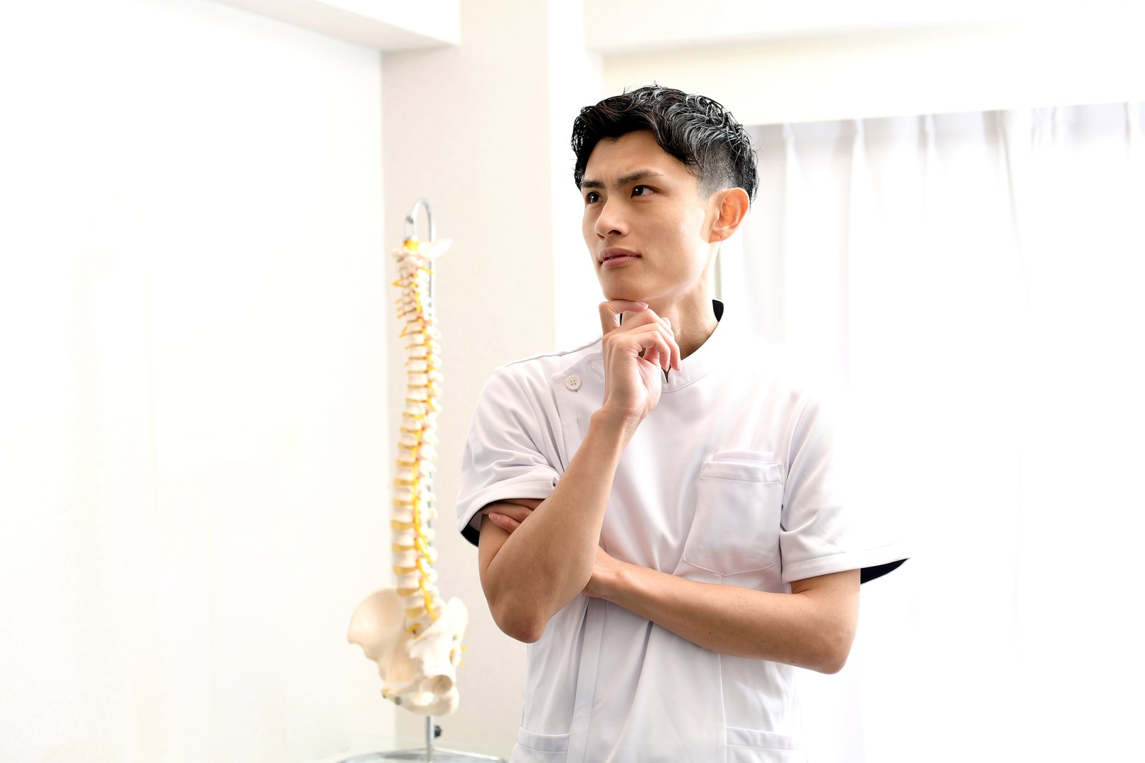 An Asian male chiropractor wearing a white coat is thinking