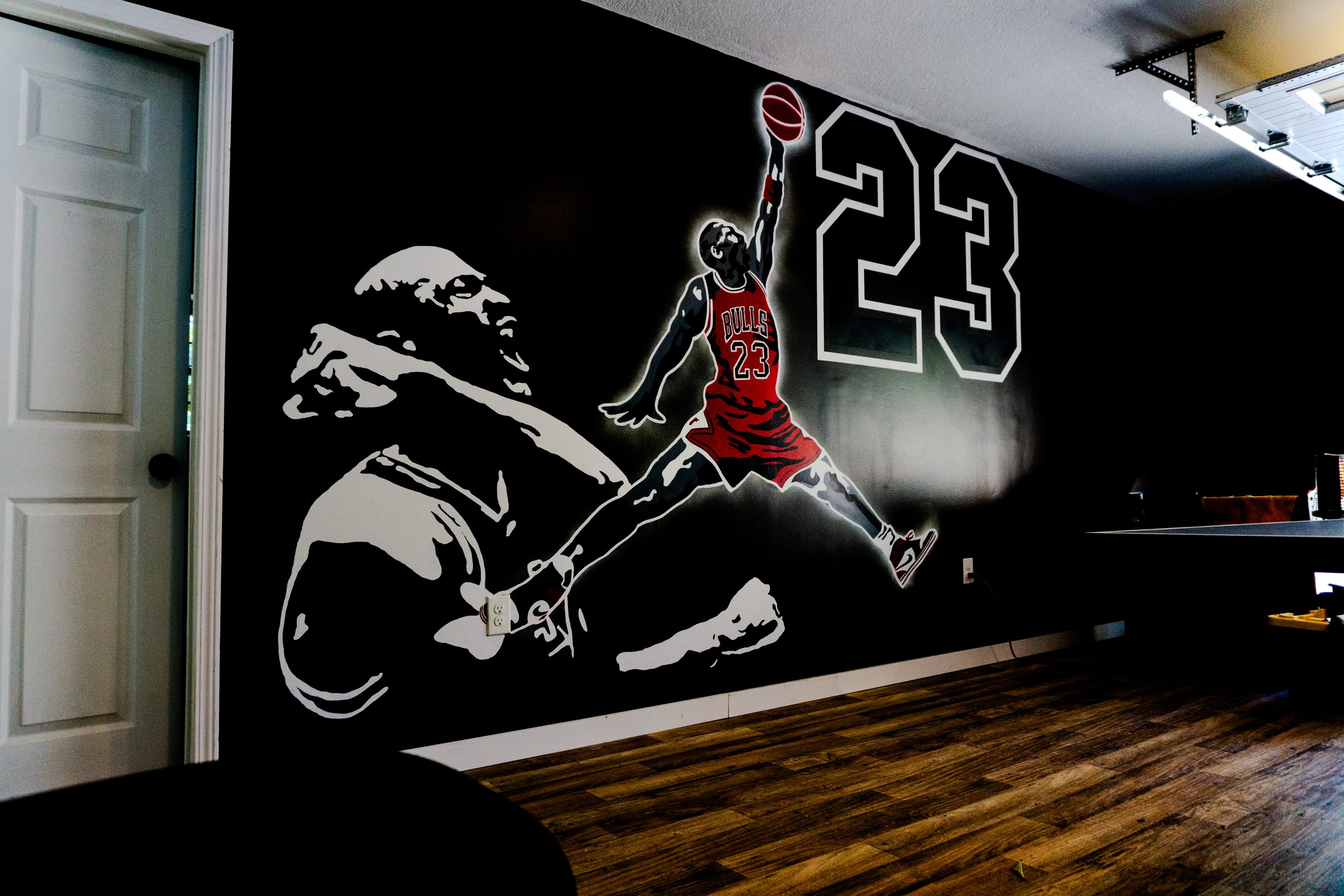 Wall mural of basketball player in mid-air next to the number 23, against a dark background, in a room with wooden flooring.