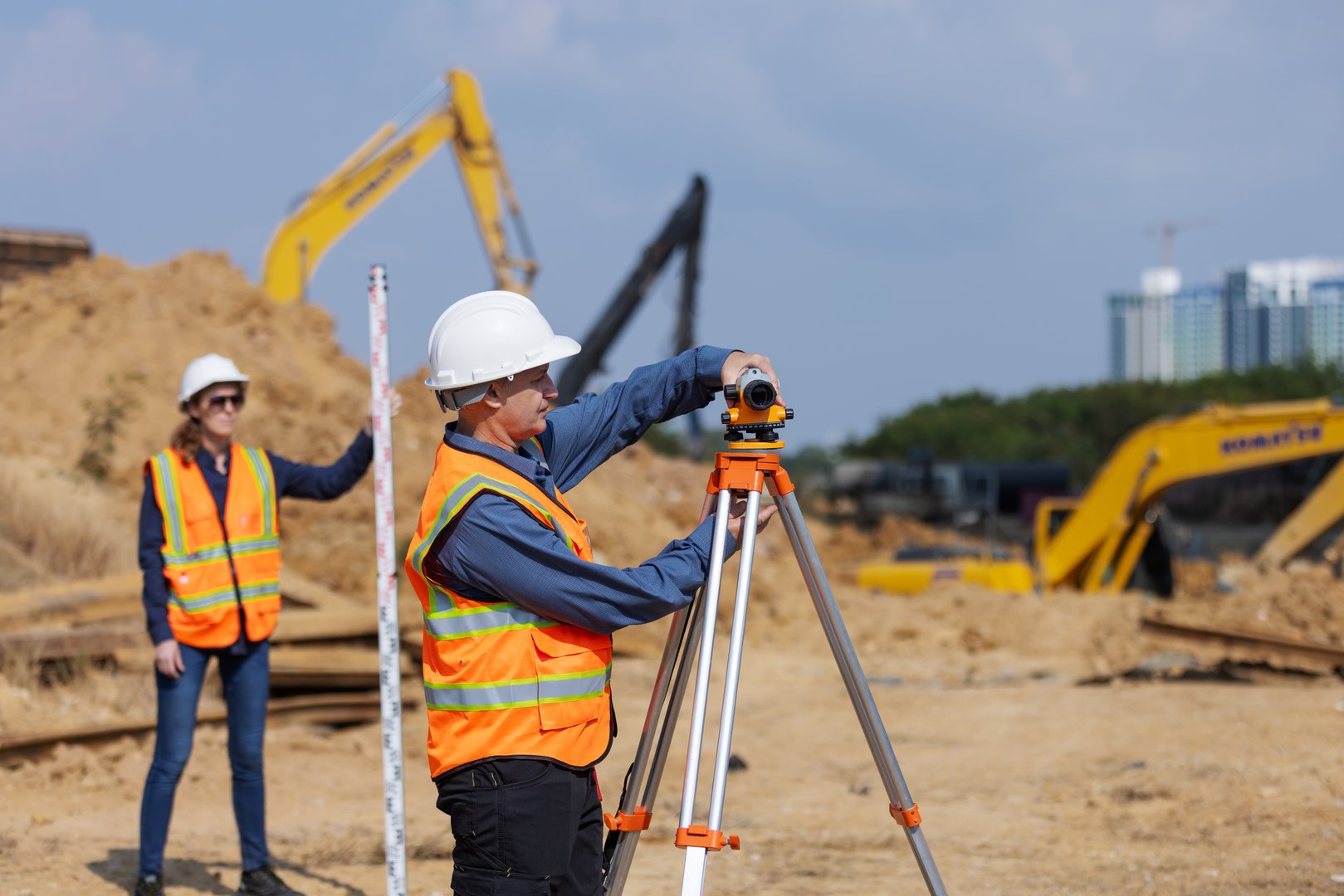 Two construction surveyors in safety gear perform land measurement at a worksite using a total station and leveling rod, surrounded by dirt piles, excavators, and construction equipment.