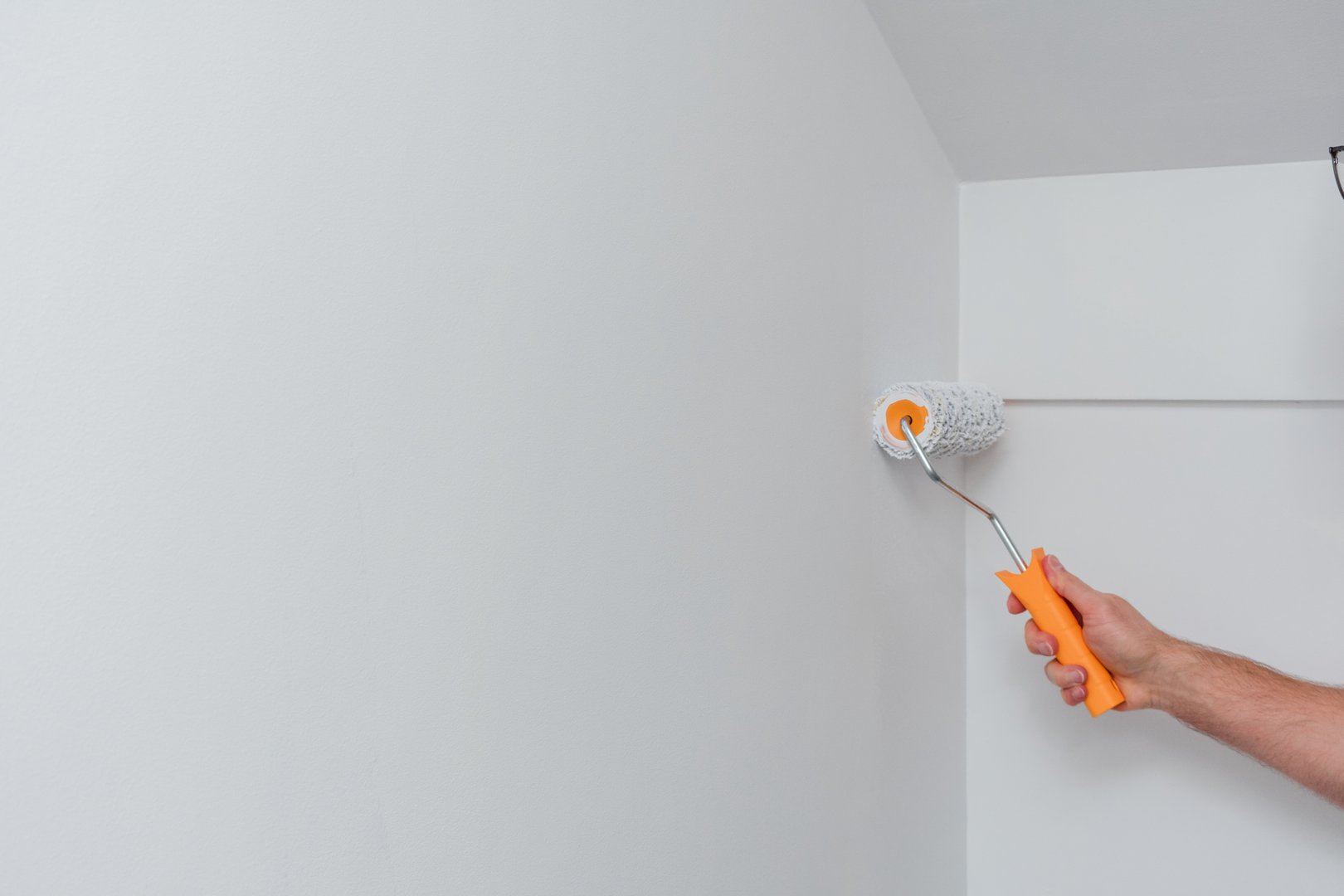 Man is painting a wall with a roller. The wall is white and the man is wearing glasses