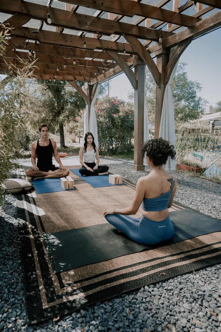 Three people doing yoga on mats under a wooden pergola, surrounded by greenery, with beams of sunlight filtering through.