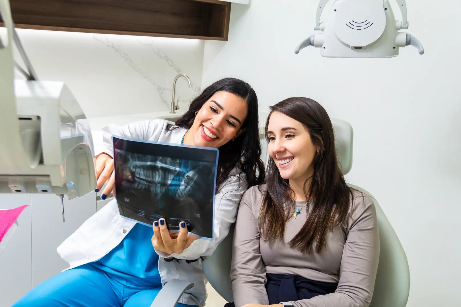 Latin female smiling dentist explaining teeth x-ray scan to caucasian woman patient sitting in a dental chair. Health care concept.