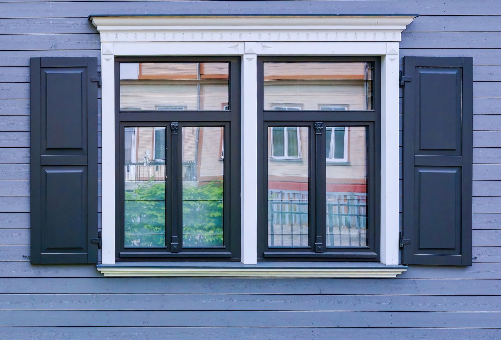 A symmetrical modern window with black shutters on a gray wooden wall, reflecting a neighboring house. Clear glass panes and minimalist exterior design create a balanced urban view.