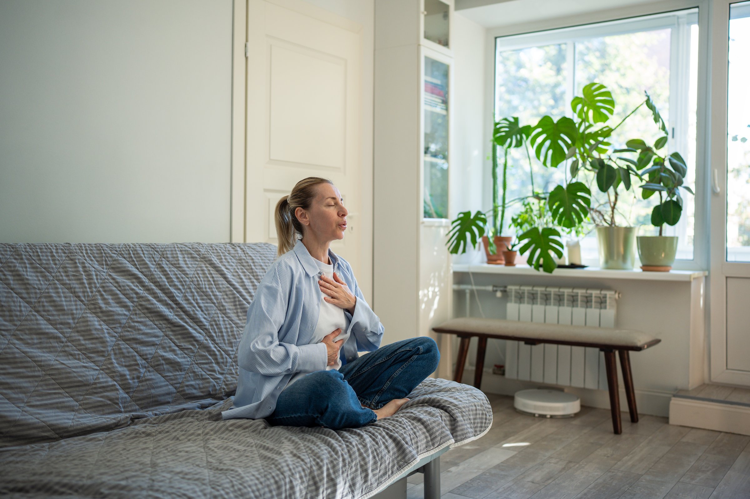Calm woman sitting in lotus pose deep breathing during meditation at home. Self-help of middle aged female stress relief in daily routine reset mind with eyes closed, placing hands on chest, abdomen.