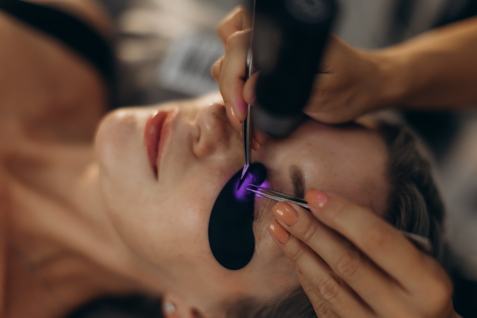 Woman getting false eyelashes applied by professional beautician. Using uv light for curing eyelash glue in a beauty salon