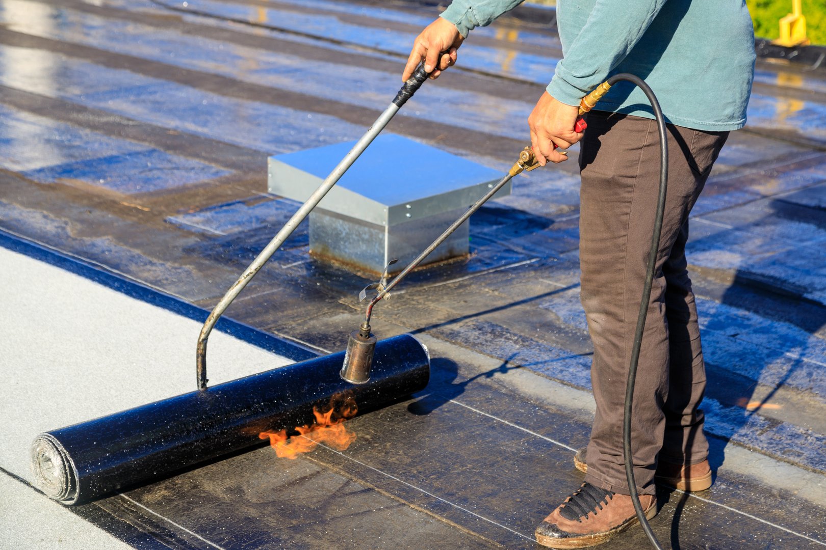 A roofer torches a membrane layer on a flat roof using a tiger torch to seal the asphalt bitumen, ensuring a durable waterproof layer.