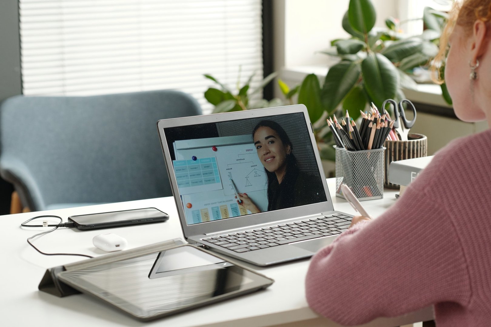 Laptop screen with young successful teacher of English grammar pointing at whiteboard with rules and explaining kinds of verbs to learner