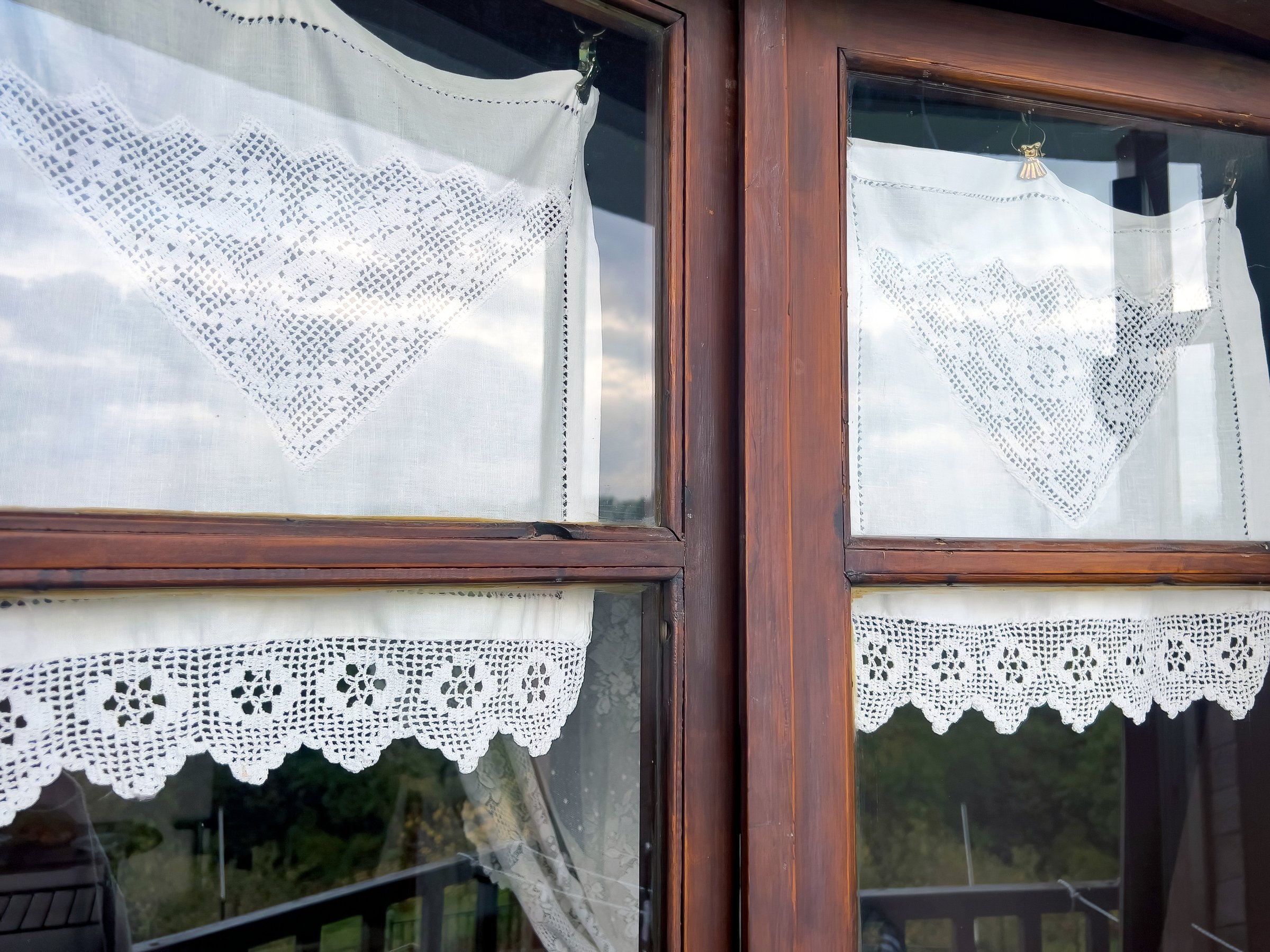 Lace curtains on wooden window reflecting sky and landscape.