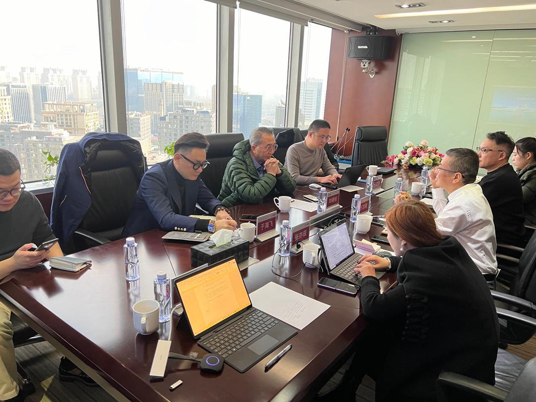People in a conference room seated around a wooden table with laptops, phones, and documents, city view through large windows.