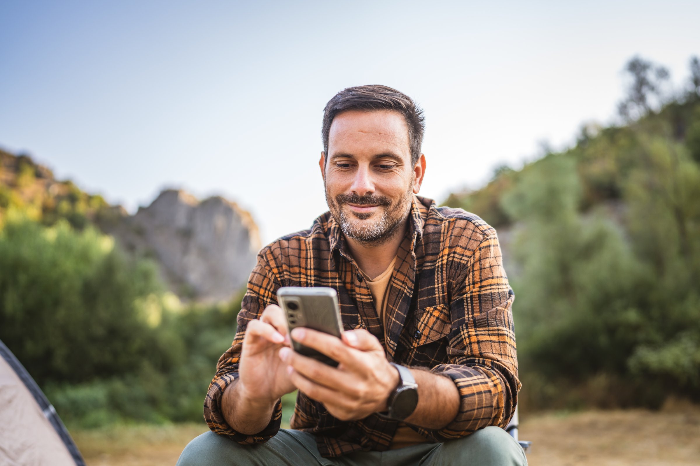 Adult caucasian man sit in front tent and use mobile phone on camping trip