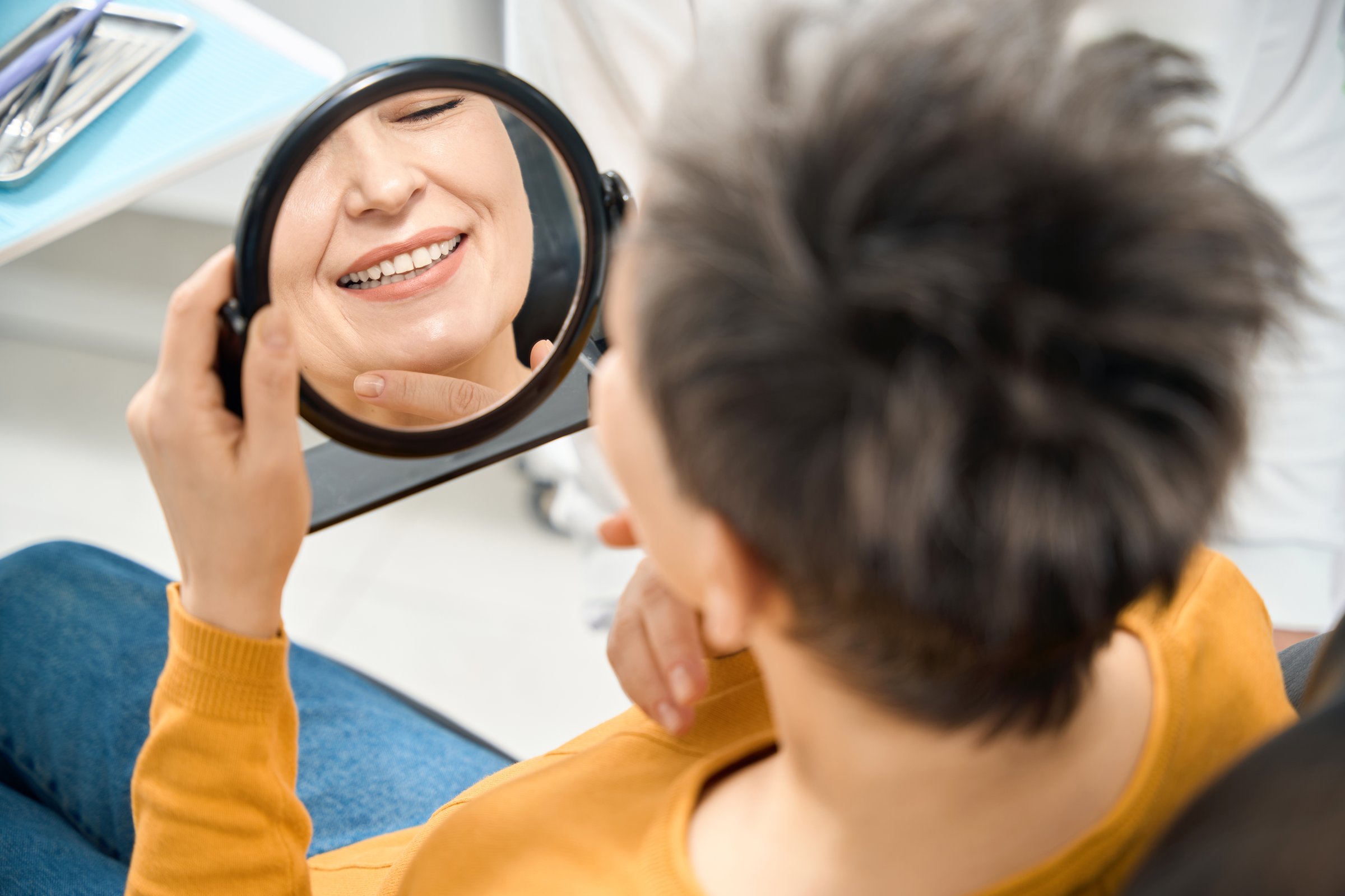 Satisfied adult woman client of aesthetic dentist checking her smile and state of teeth after professional hygienic cleaning session looking at reflection in the mirror
