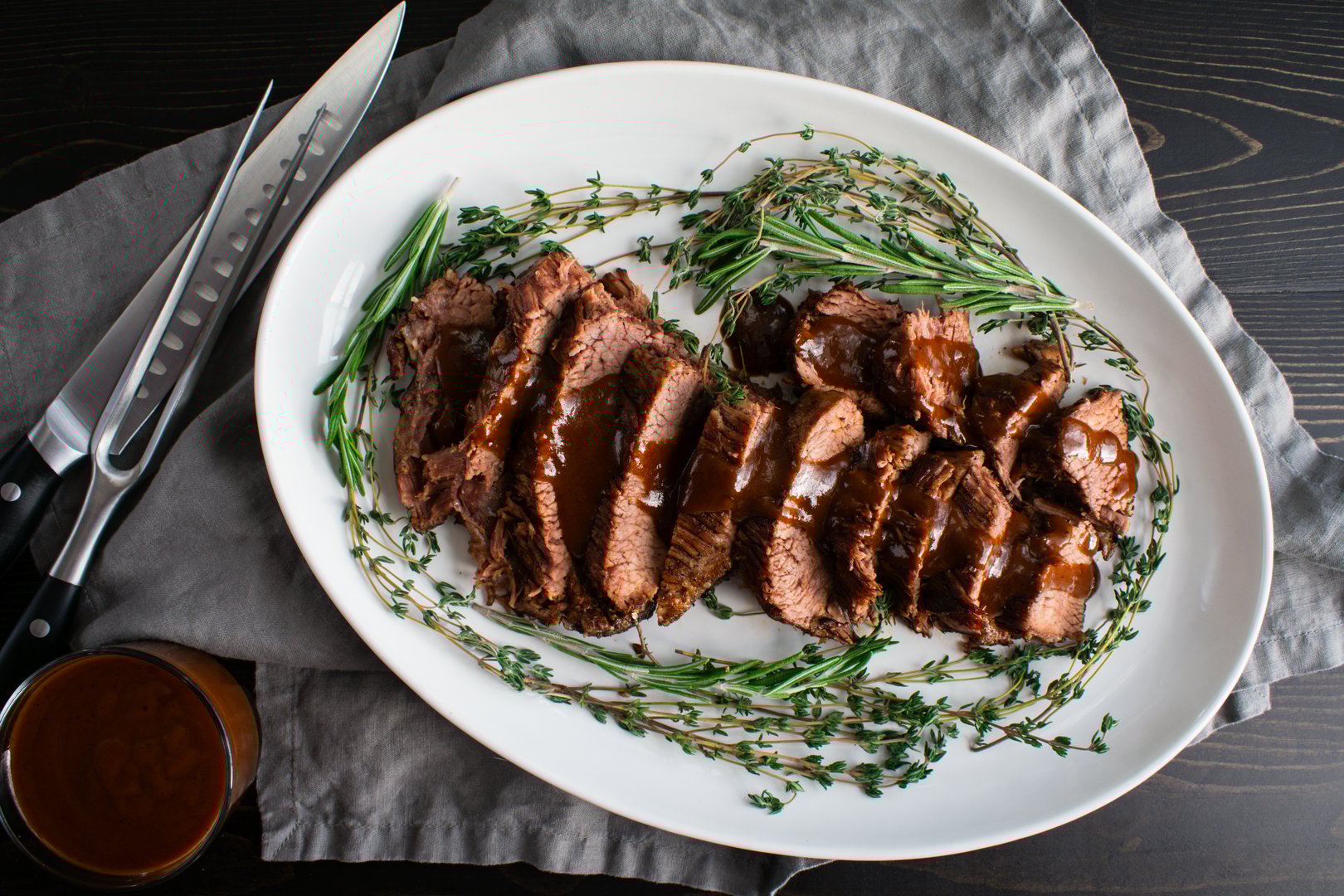 Slices of roast beef with sauce surrounded with sprigs of thyme and rosemary on a ceramic plate