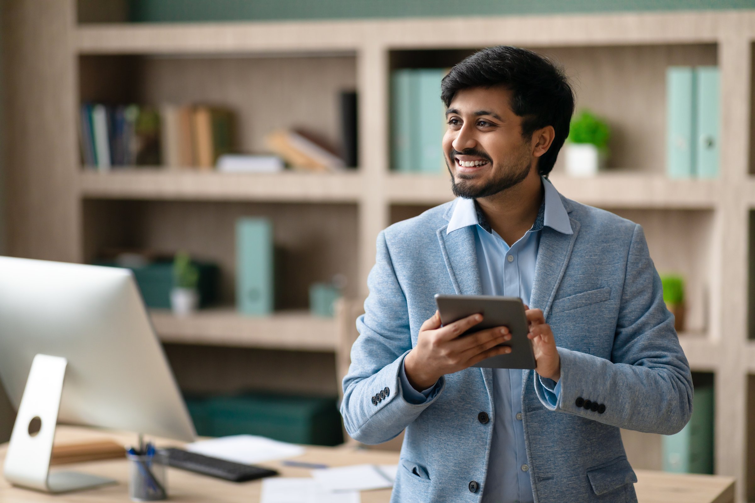 Digital Professional. Happy Indian programmer man using tablet computer standing in modern office with desktop computer, surfing web during work day, smiling looking away. Tech and business