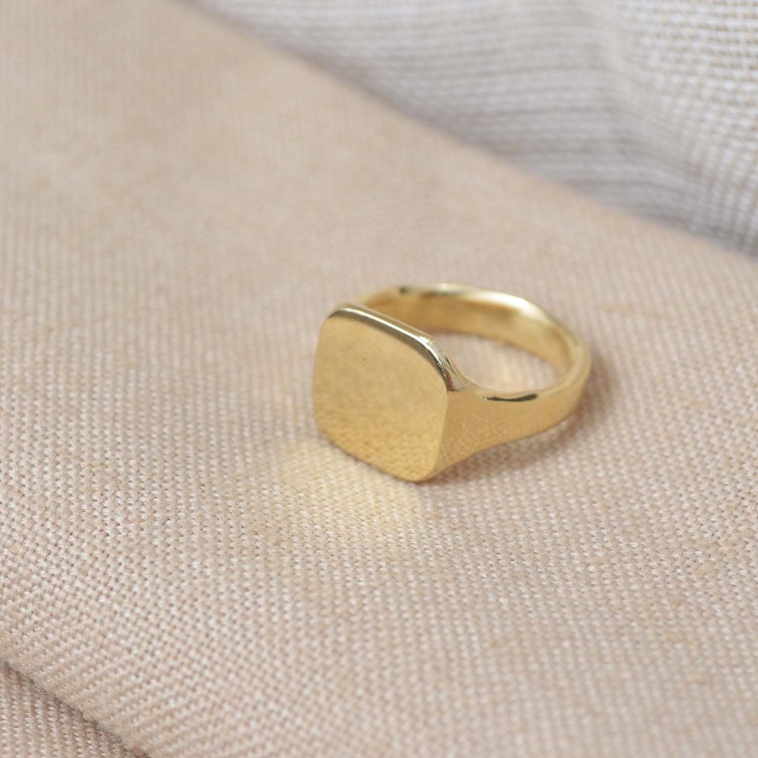 Gold signet ring with a textured square face on a beige textile surface.