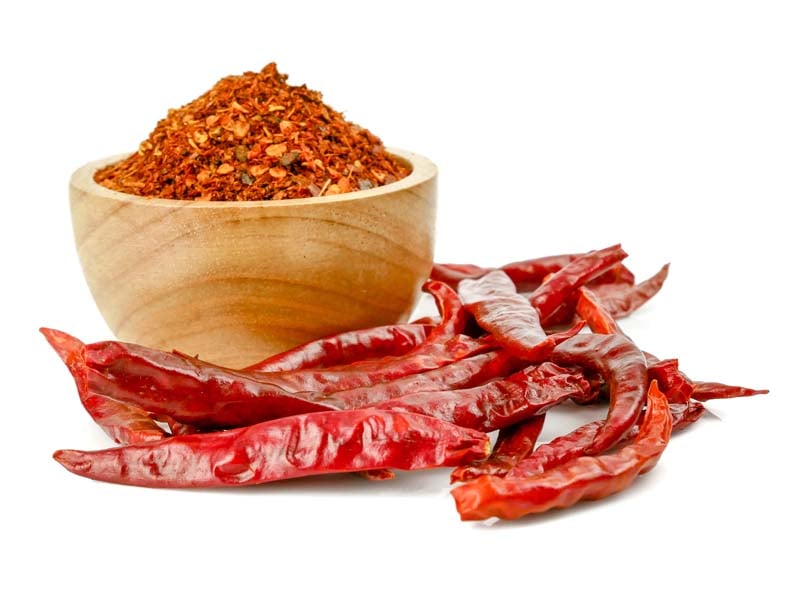A wooden bowl of chili powder next to several whole dried red chilies, isolated on a white background.