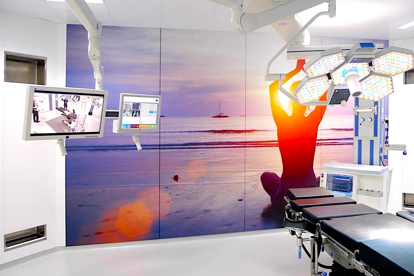 A modern operating room with a beach sunset mural, surgical lights, and monitors displaying medical images.