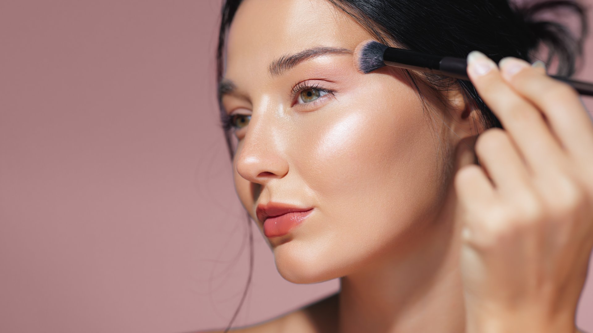 Detailed close-up of a young woman applying shimmering highlighter on her cheekbone with a makeup brush, highlighting her radiant and flawless skin. Ideal for beauty, makeup, and skincare promotions.