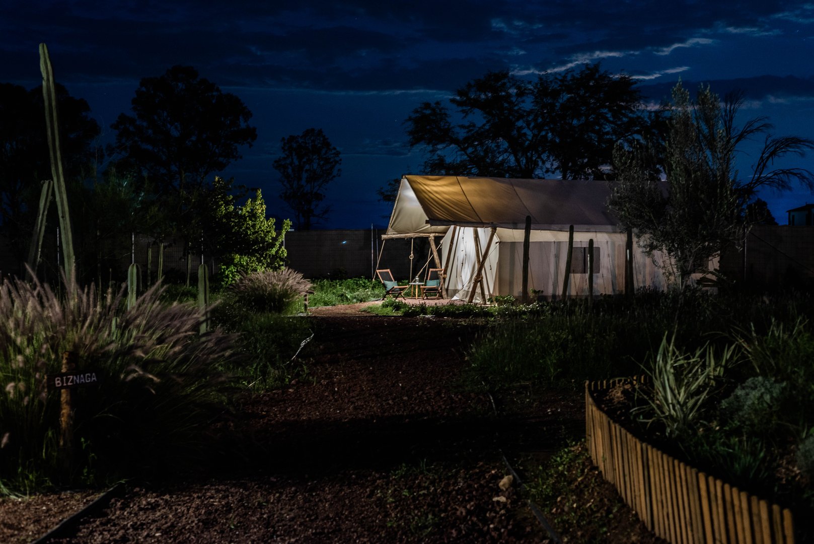 A dimly lit tent with a small porch surrounded by plants and trees at night, under a cloudy sky.