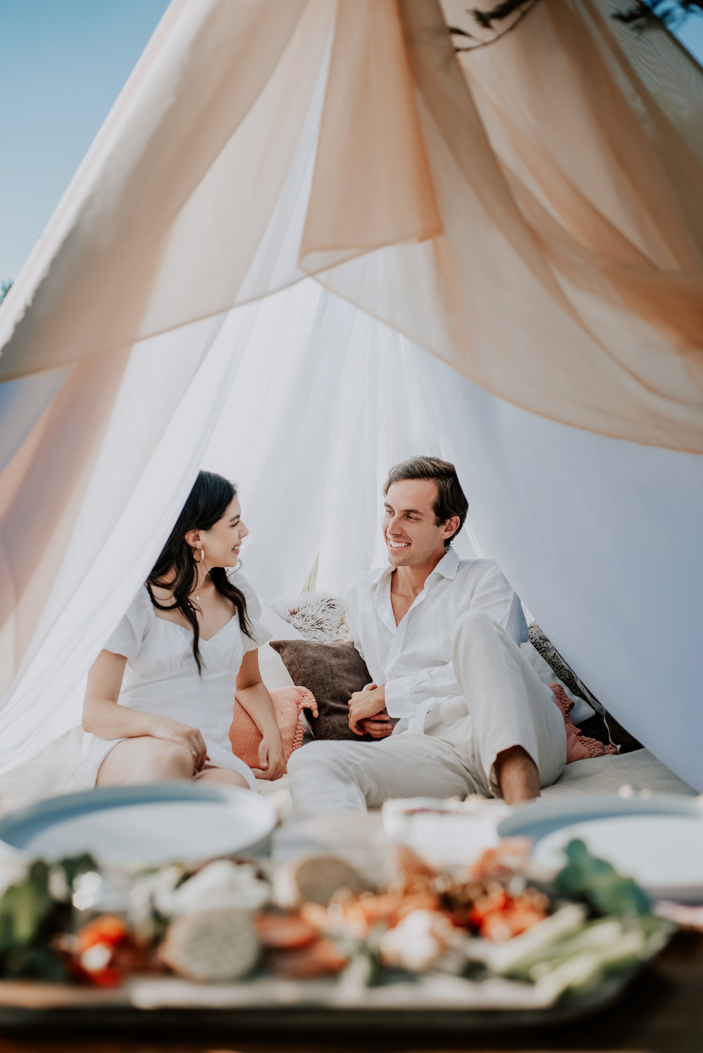 Couple sitting in a cozy tent with soft pillows, smiling at each other. A picnic setup with plates is in the foreground.