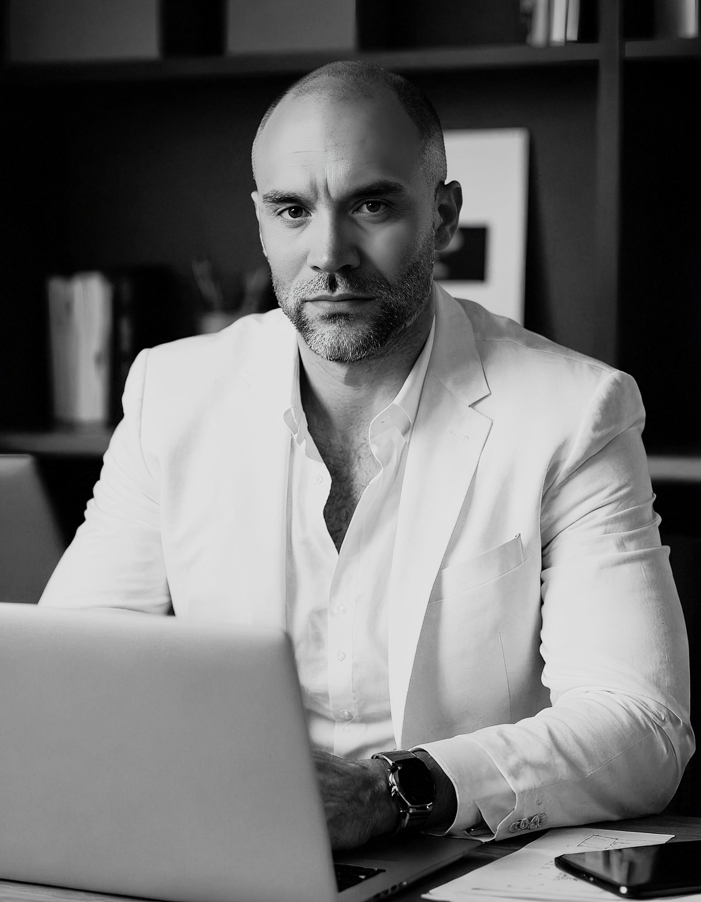 Man in a white suit working on a laptop at a desk, with shelves and books in the background.