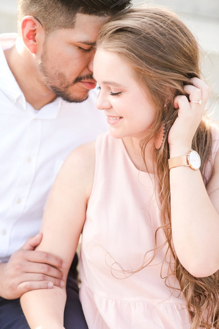 A couple sitting close together, smiling, with the man gently kissing the woman's head. She touches her hair, wearing a watch.