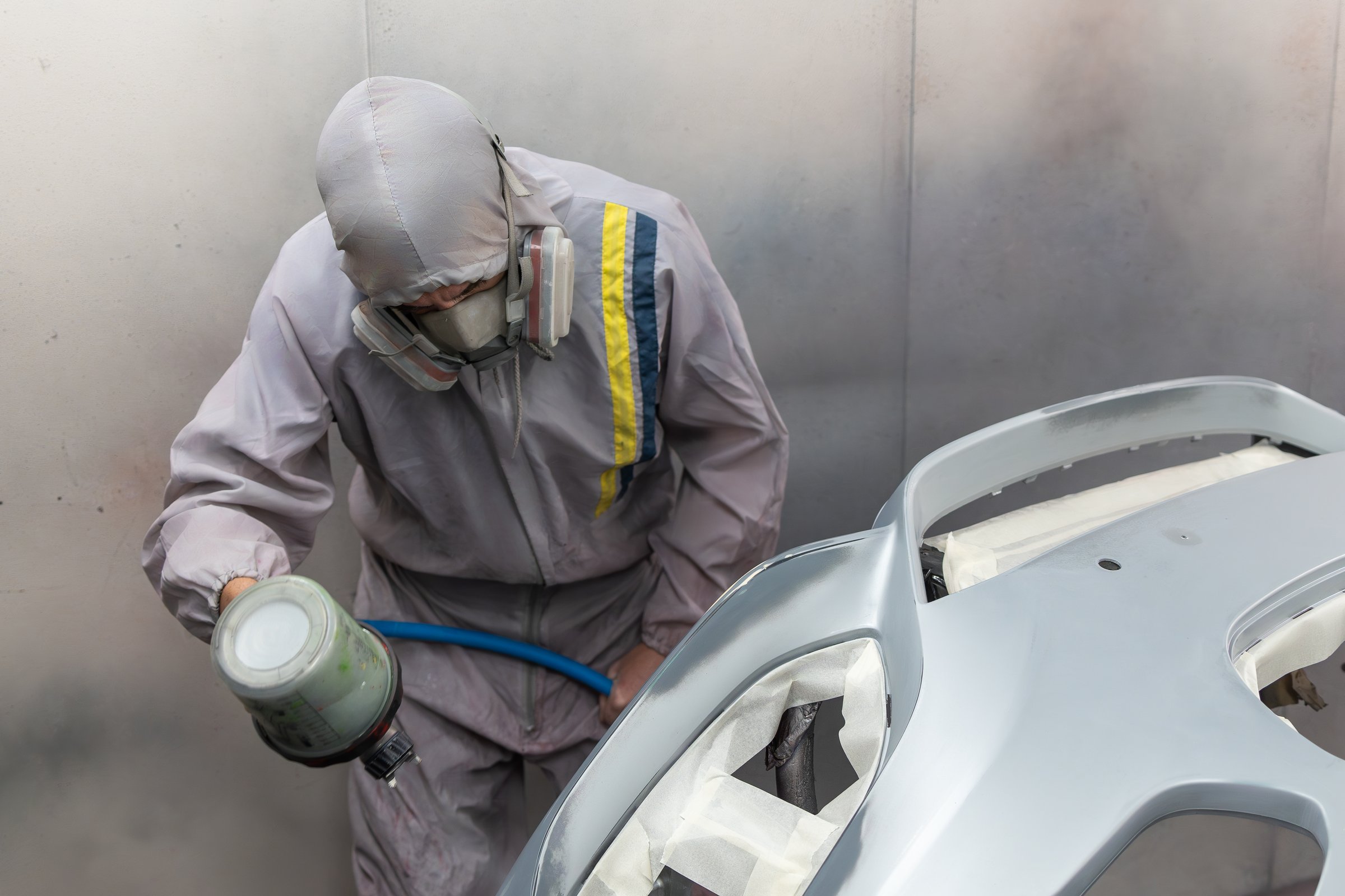 Painter in full protective suit focuses intently on spraying a car hood with precision in a professional paint booth.