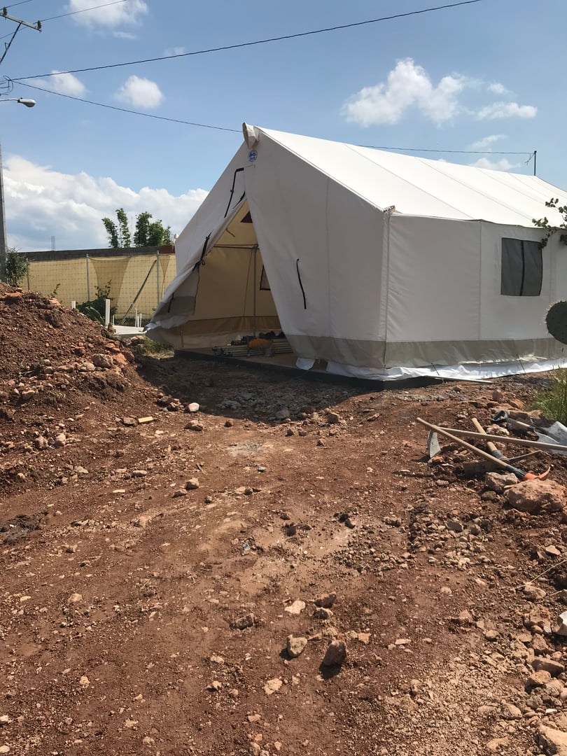 Large white tent on a dirt and rocky ground with a clear sky, partially open entrance, and construction tools nearby.