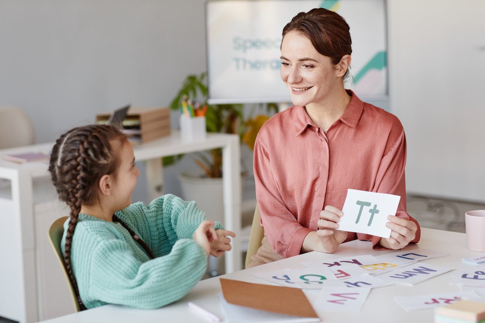 Young teacher showing the card with English letter while teaching the little girl at the table in the room