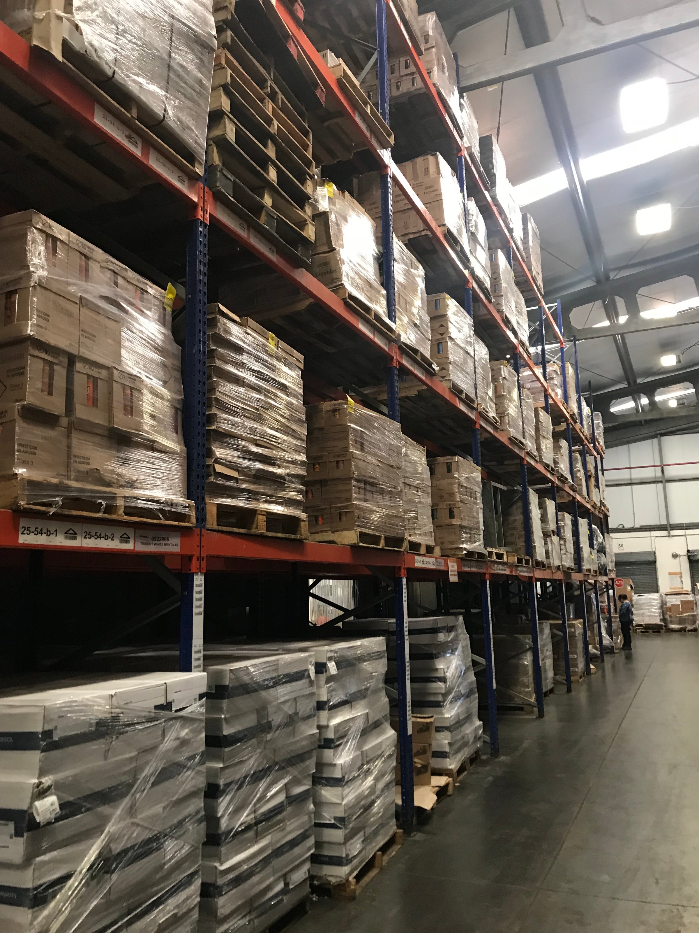 A warehouse with tall shelves filled with neatly stacked pallets of boxes and goods, under industrial lighting.