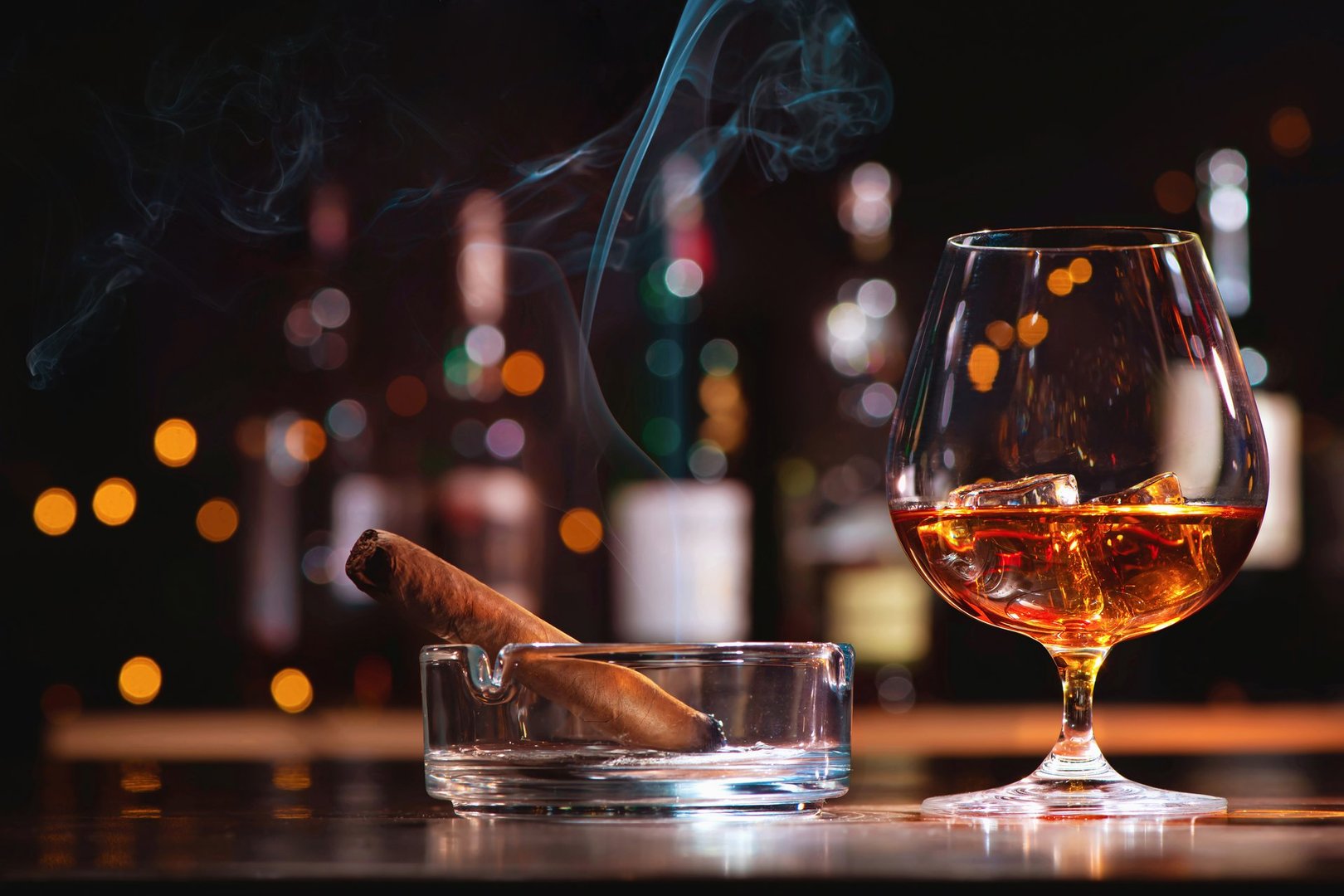 Glass of whiskey or cognac with ice cubes and smoking cigar and on bar counter