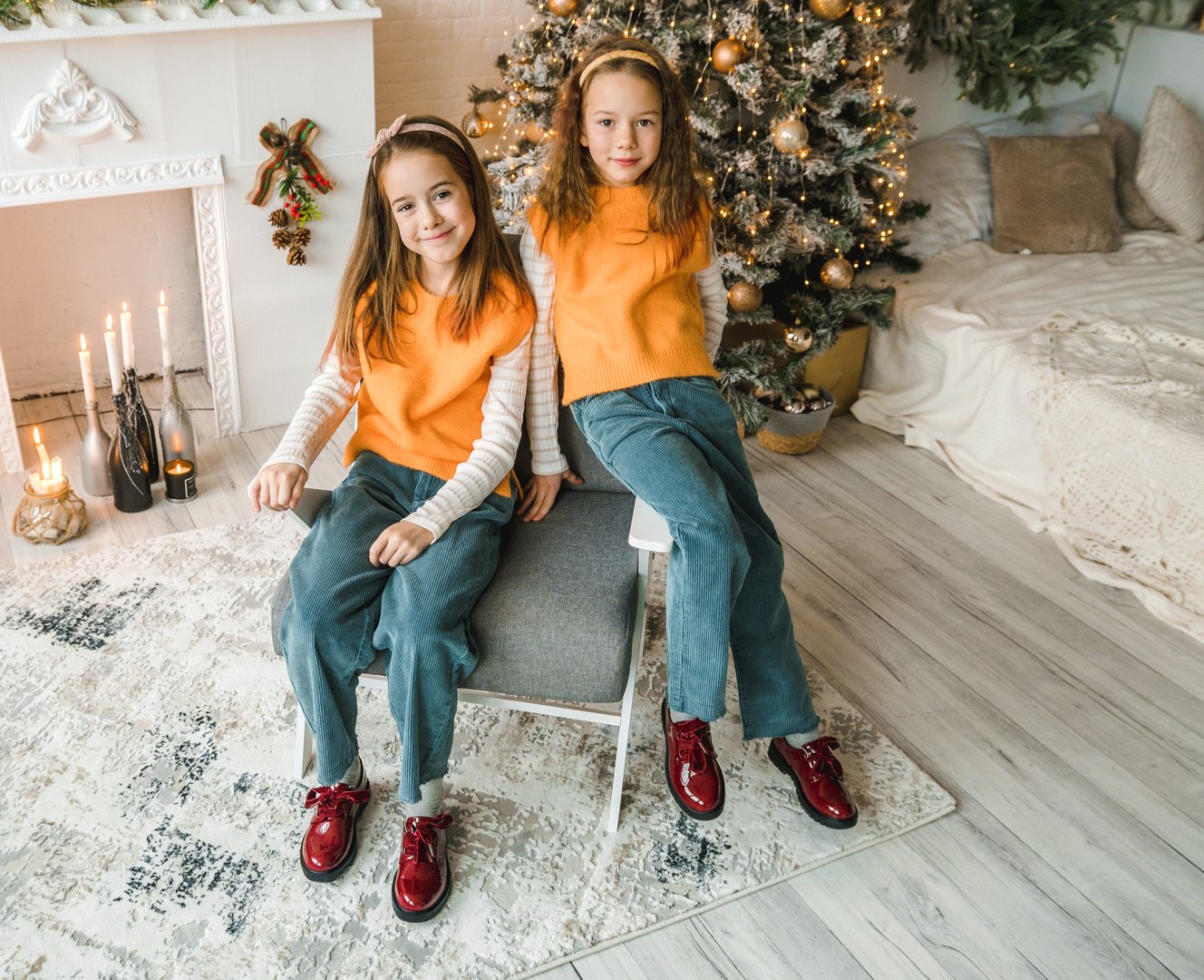 Two adorable twin girls, dressed in matching orange sweaters and denim pants, pose for a Christmas portrait. They are sitting in a chair, wearing bright red shoes, in a room decorated for the holidays.