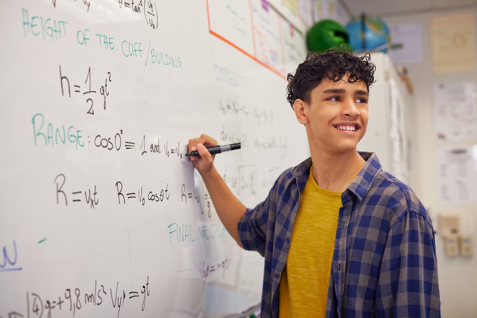 Multiethnic teenager student smiling while writing equation on whiteboard. Latin teen solving physics problems during lesson in classroom. Confident guy participates in a math lesson.