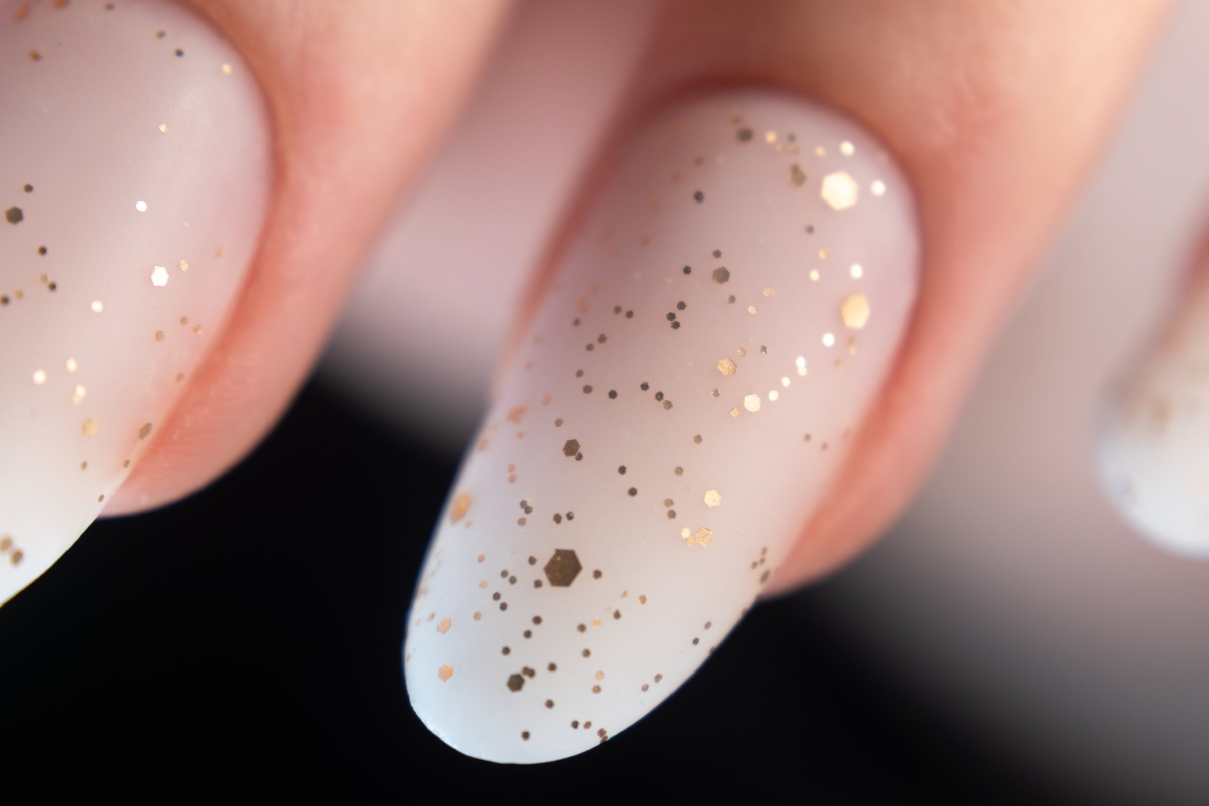 A close-up of fingernails featuring a white polish base adorned with gold glitter. The glitter particles vary in size and shape, creating a speckled effect across the nails. The nails are well-manicured, with an oval shape, and the gold elements add a touch of sparkle and elegance. The image focuses on the aesthetic detail and texture of the nail design.