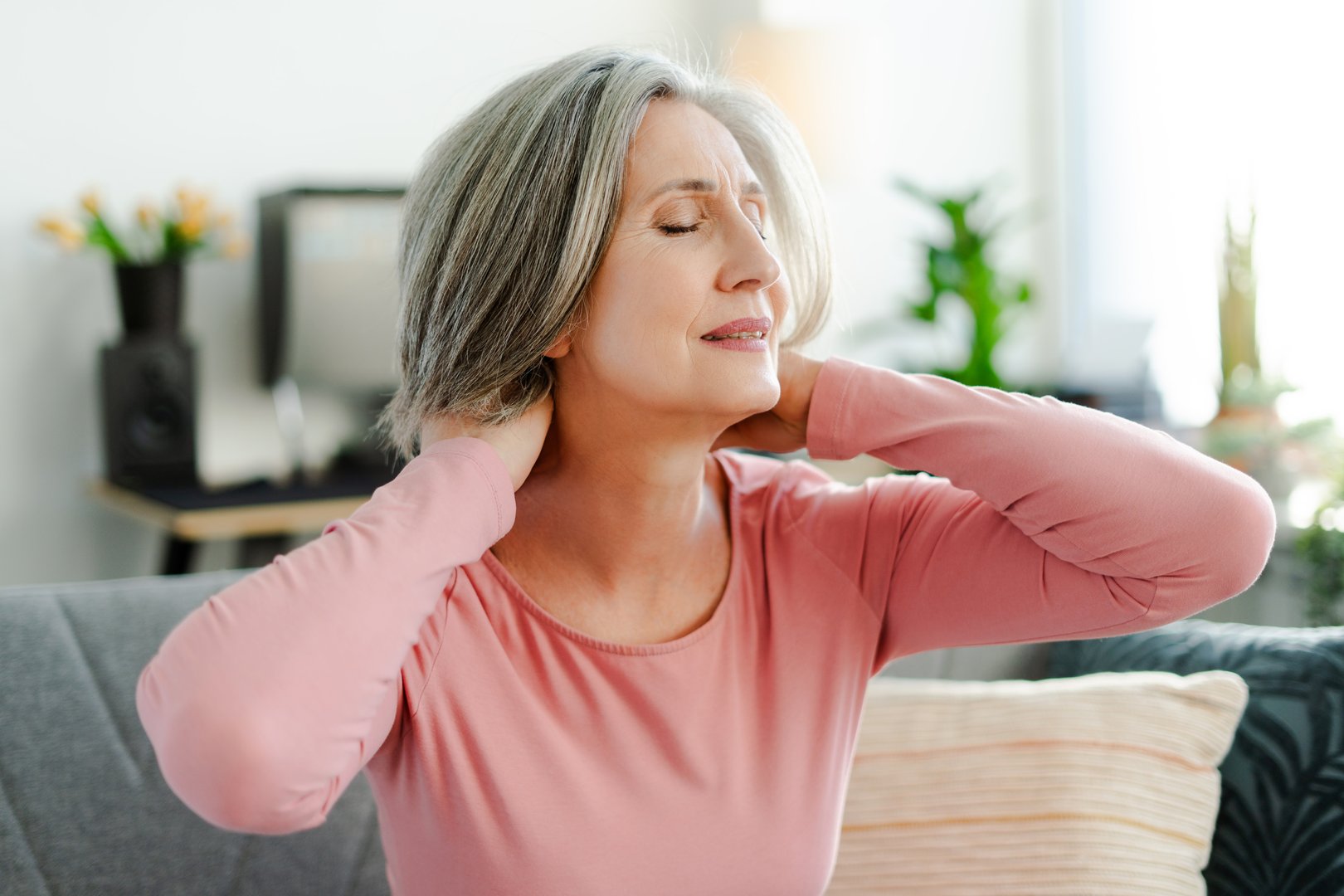 Unhappy senior woman stretching arms, doing exercises, having neck pain sitting on comfortable sofa relaxing at home. Sad female, pensioner taking break, resting