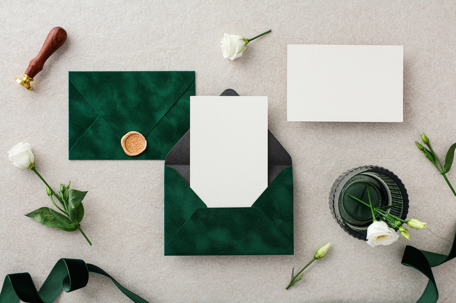 Wedding stationery set on stone desk top view. Blank paper card mockup, dark green envelopes with wax seal stamp, green ribbons, flowers.