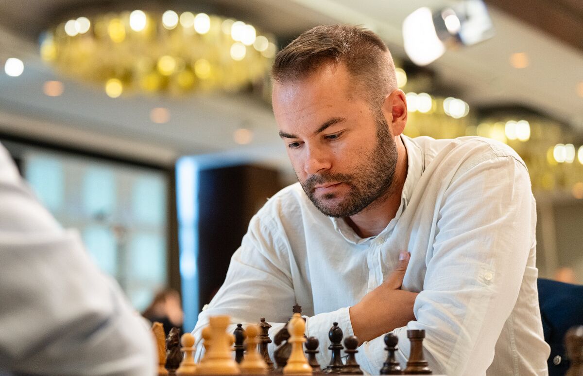 Man concentrating on a chess game in an elegant room, wearing a light shirt, with blurred chandeliers in the background.