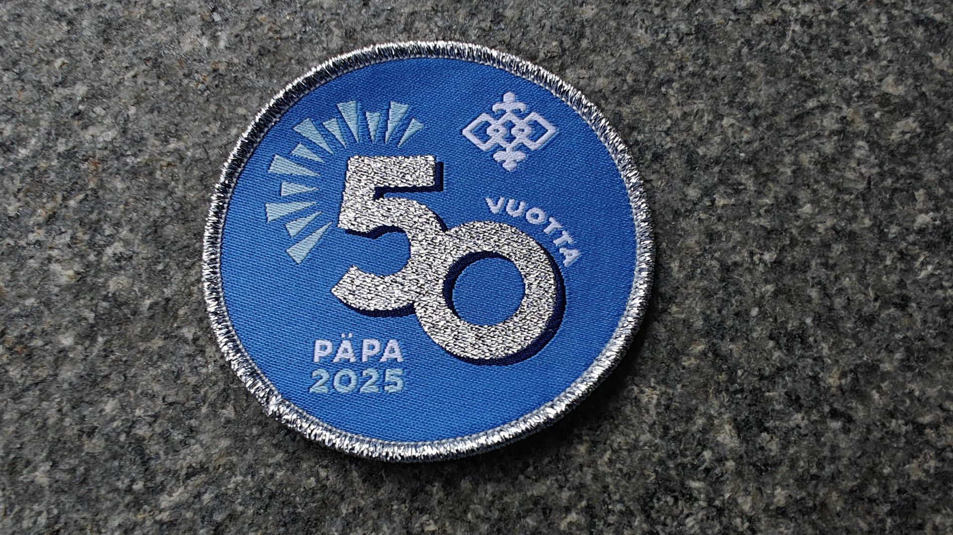 Blue circular patch with glittery number 50, text vuotta 2025 and Päpa, featuring a pattern on a stone surface.