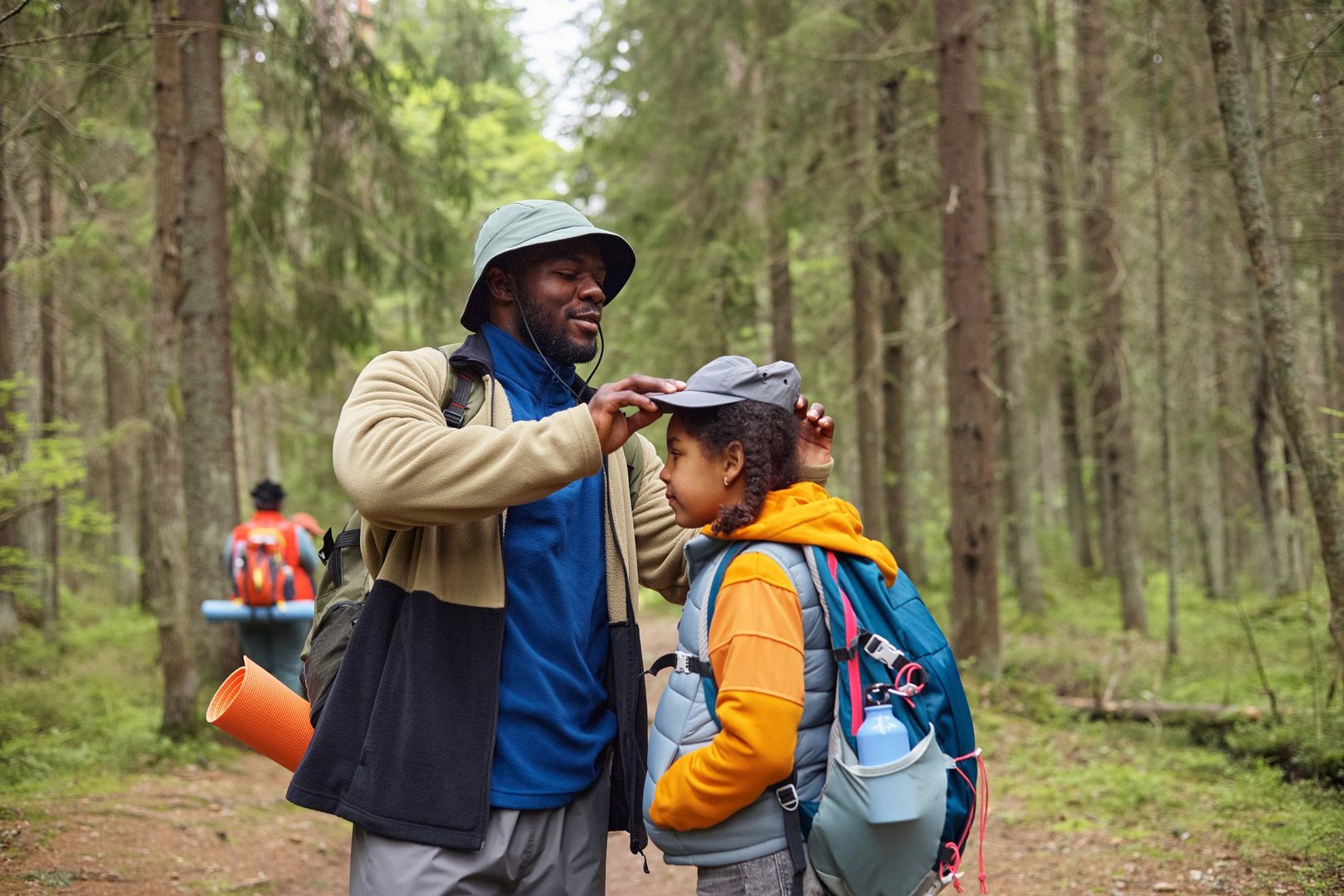 Black middle aged man adjusting cap of Black child while hiking in forest, both carrying backpacks and outdoor gear, another with backpack and mat walking ahead among trees
