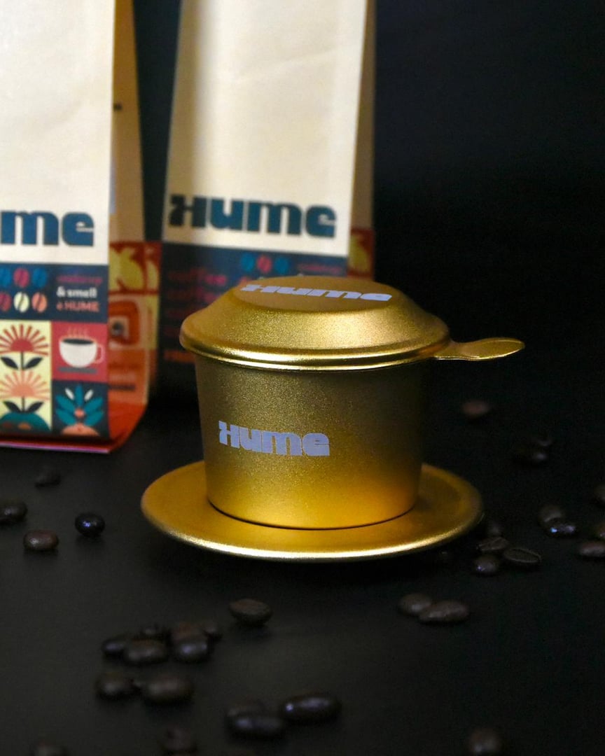 A gold coffee dripper with "Hume" branding, surrounded by coffee beans, next to packaged coffee on a dark background.